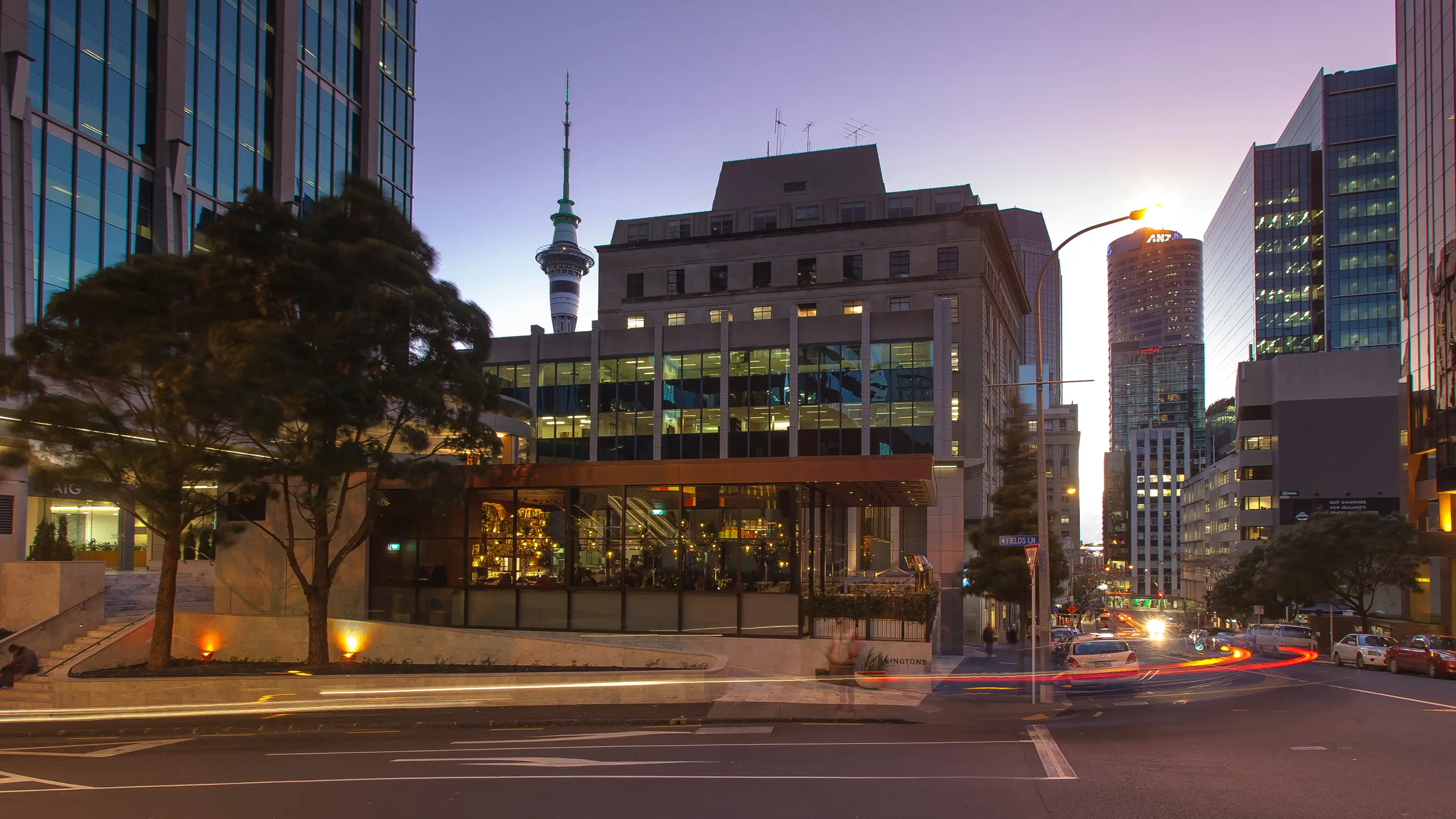 Pilkingtons: the glass restaurant pavilion lit up at dusk on an Auckland CBD corner, Sky Tower behind